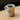 White mug with coffee on a yellow surface, featuring 'The Grounds Roastery' logo. 